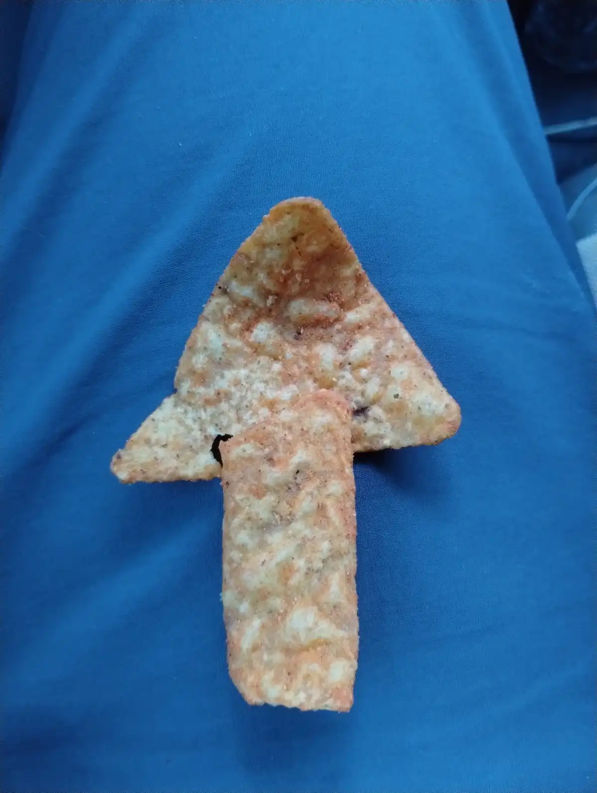 A single, broken, seasoned tortilla chip is arranged to resemble an arrow pointing upwards, resting on a blue fabric…