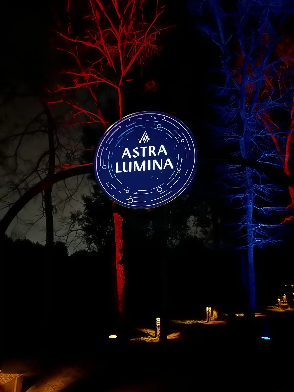 A circular blue sign with white text reading 'ASTRA LUMINA' is illuminated at night, surrounded by trees bathed in red and…