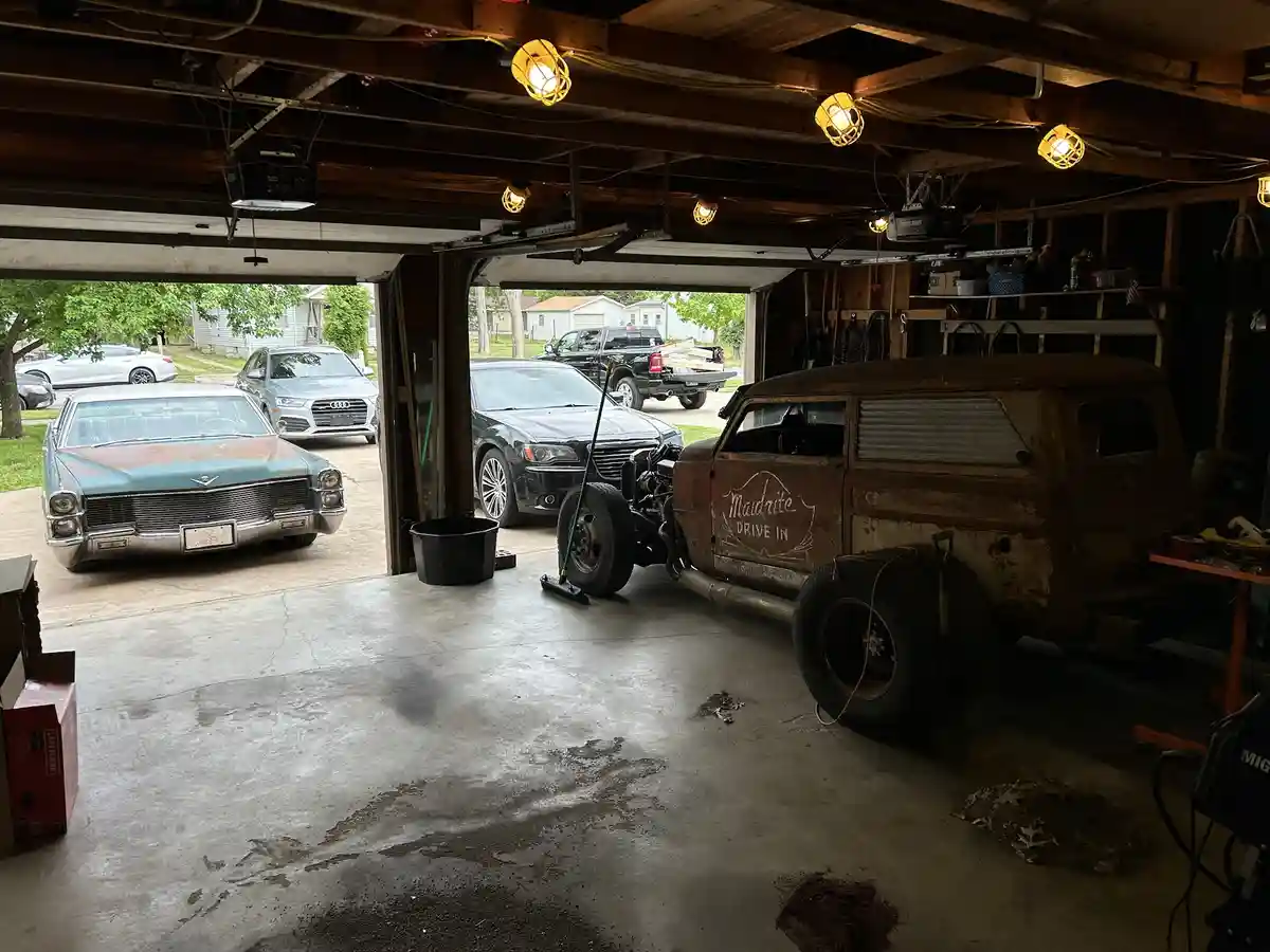 A cluttered garage contains a rusty, stripped-down vintage station wagon with 'Maidrite Drive In' painted on its side…