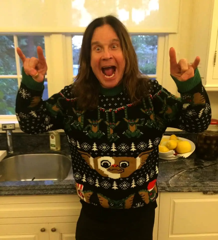 A man with long brown hair stands in a kitchen wearing a black Christmas sweater featuring a large Gremlins character and…