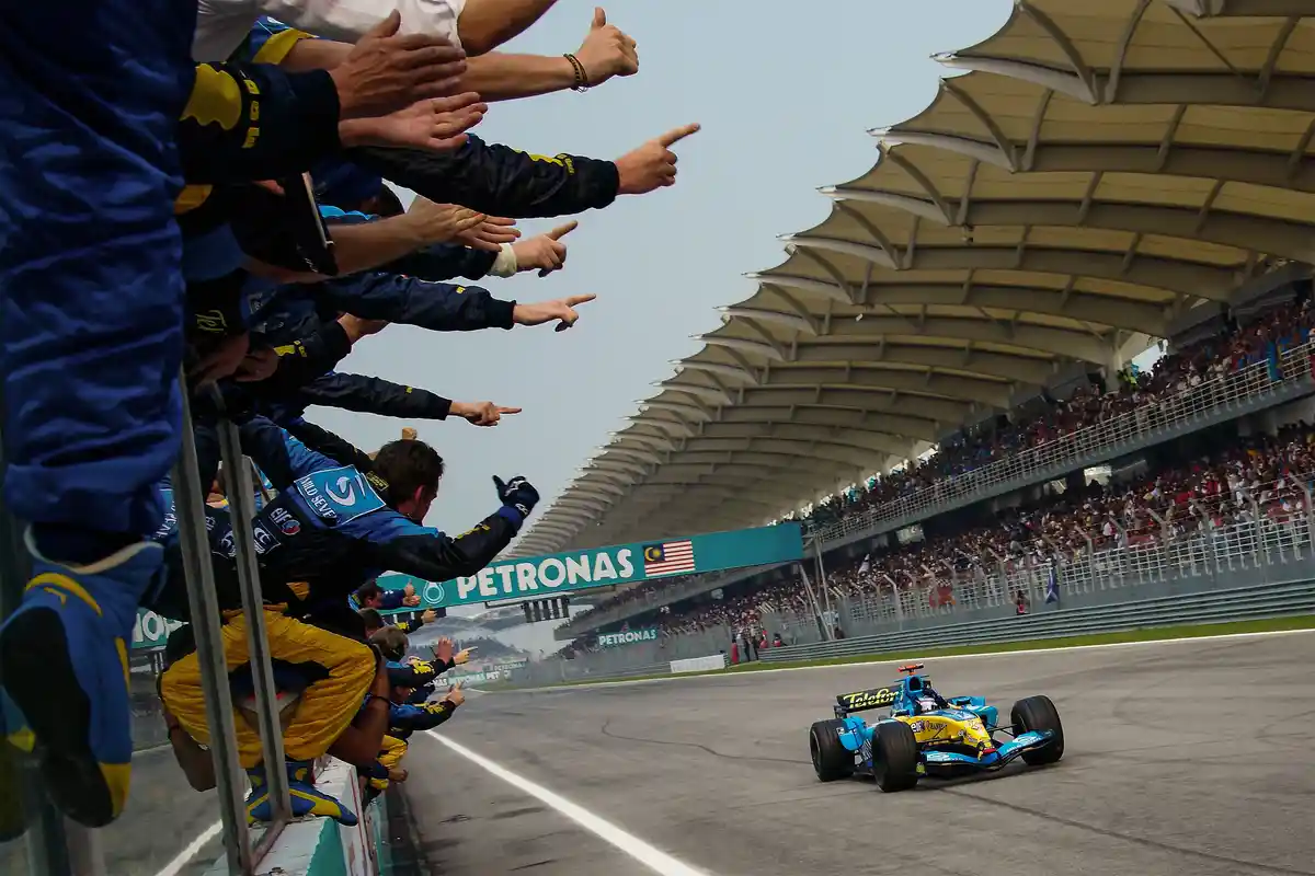A blue and yellow Formula 1 race car speeds along a track as a crowd of team members in matching blue and yellow uniforms…