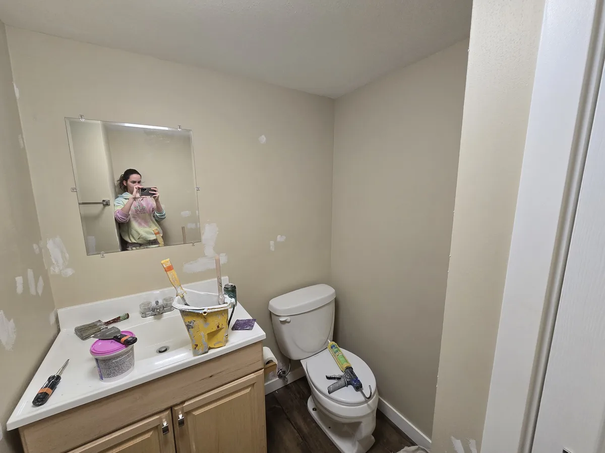 A person takes a mirror selfie in a bathroom under renovation, with visible drywall patches on the walls and tools like a…