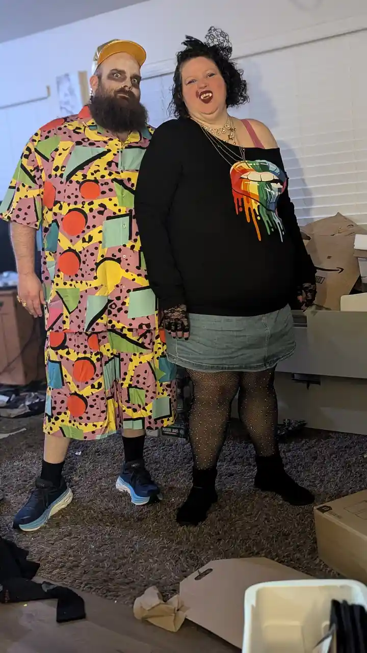 Two people pose indoors, one wearing a colorful, geometric-patterned outfit with a yellow cap and dark makeup, the other in…
