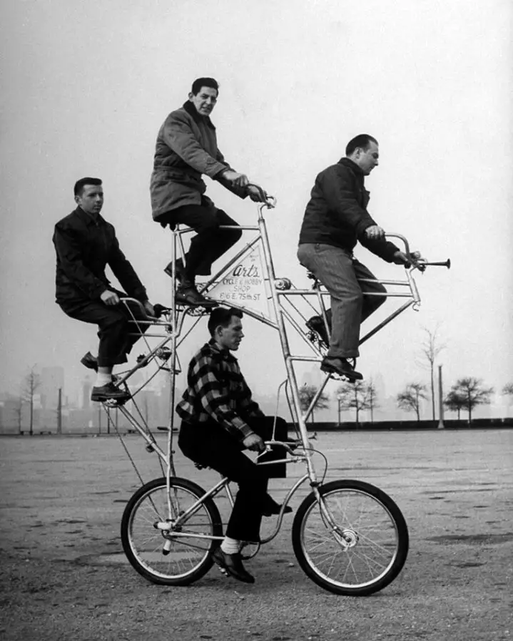 a black-and-white photograph shows four men riding a large, custom-built, multi-person bicycle in an open paved area. The…