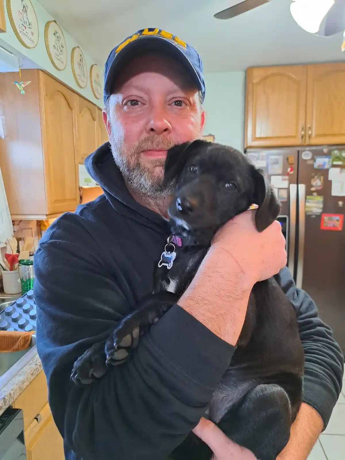 A man in a blue cap and black hoodie holds a small black puppy in a kitchen, looking directly at the camera with a neutral…