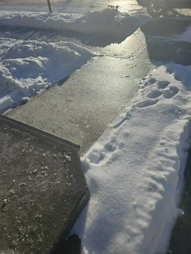 A close-up photo shows a sidewalk with melting snow and ice, where a large, jagged snowbank on the right has been shaped by…