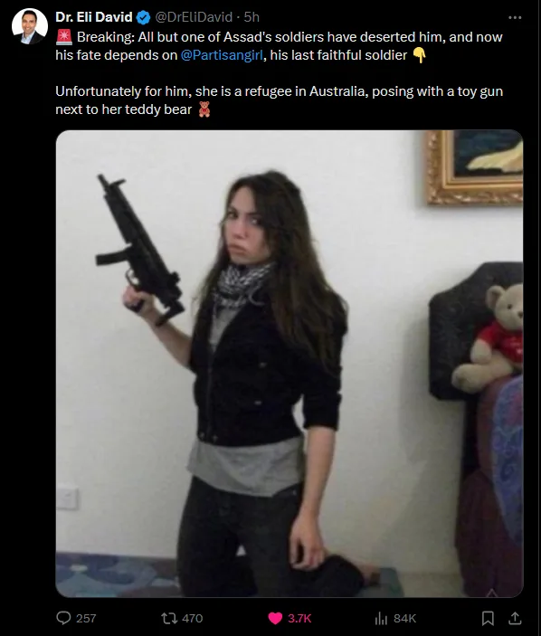 A woman with long dark hair, wearing a black jacket over a grey shirt and dark pants, kneels while holding a black toy gun…