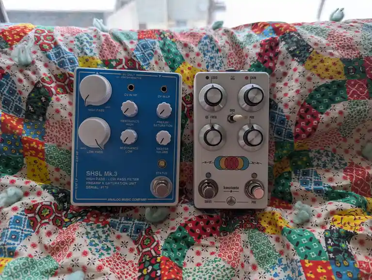 Two analog guitar effects pedals sit side-by-side on a colorful patchwork quilt. The blue pedal on the left is labeled 'SHSL…