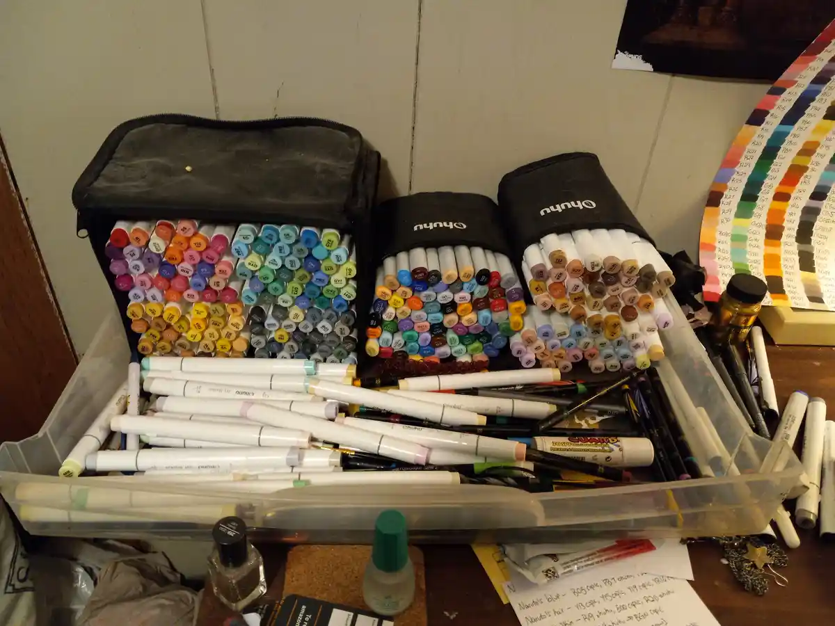 A cluttered artist's desk shows three open black zippered cases filled with numerous colorful Ohuhu art markers arranged by…