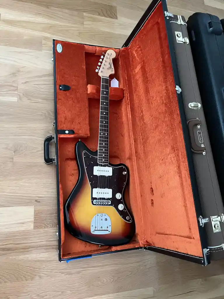 A sunburst Fender electric guitar rests inside an open, orange-lined hardshell case on a light wood floor. The case is…