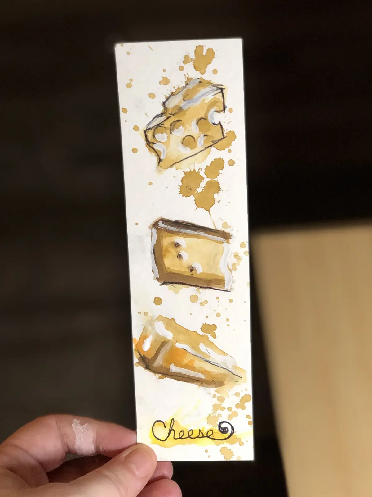A hand holds a vertically oriented white bookmark with a watercolor illustration of three different types of cheese: a wedge…