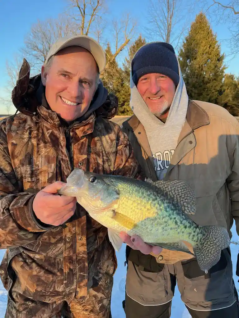 Two smiling men in winter clothing stand on snow-covered ground holding a large, speckled fish, likely a crappie, with one…