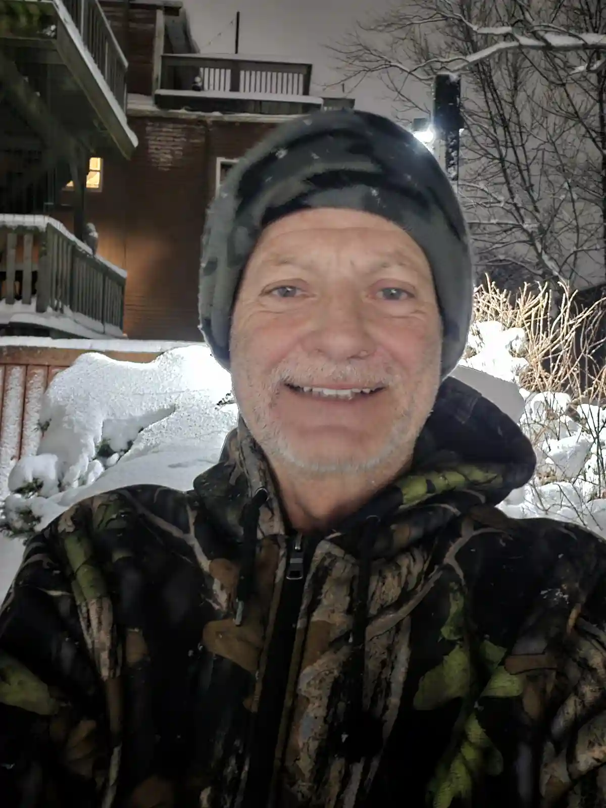 A man in a camouflage jacket and beanie smiles at the camera while standing outside in a snowy, nighttime setting with…