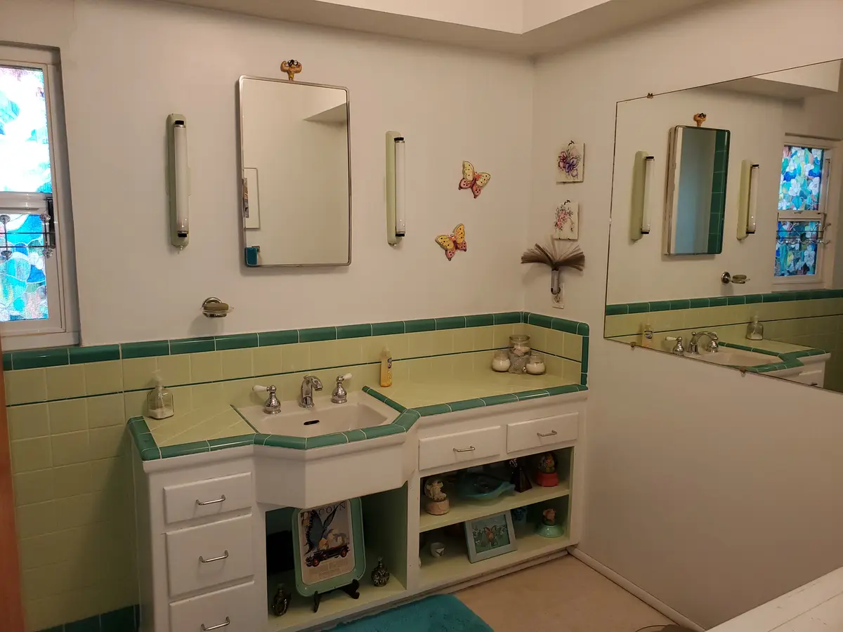A vintage-style bathroom features a white vanity with a green-tiled countertop and backsplash, two wall-mounted mirrors, and…