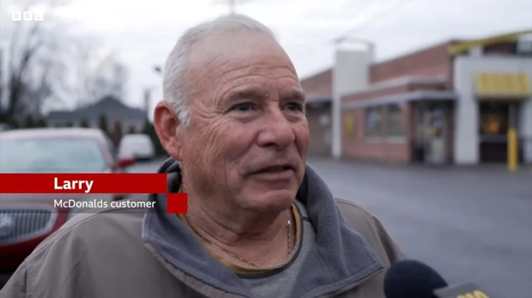 An older man with gray hair, wearing a gray jacket, is being interviewed outdoors in front of a McDonald's restaurant. A red…