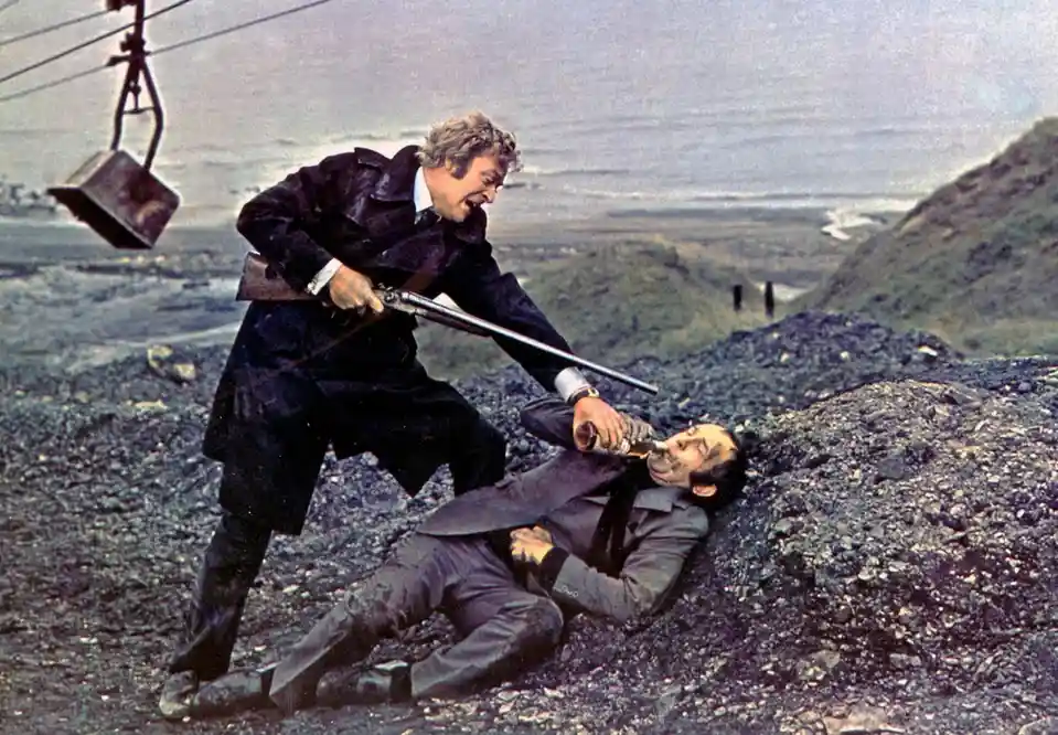 A man in a dark coat stands over another man lying on gravel, holding a rifle and forcing a bottle to the other man's mouth…