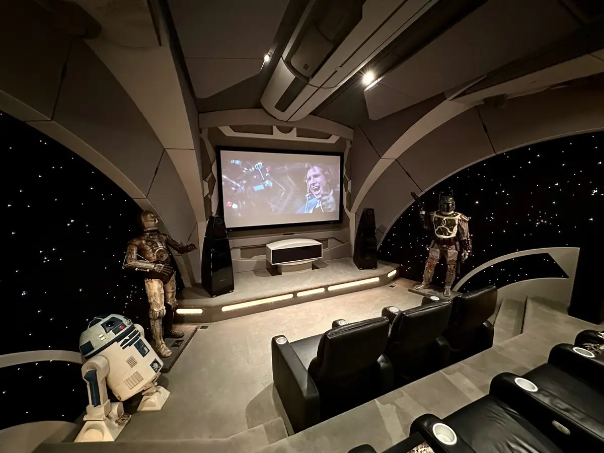 A high-end home theater room with a curved screen showing a movie, flanked by life-sized statues of Star Wars characters…