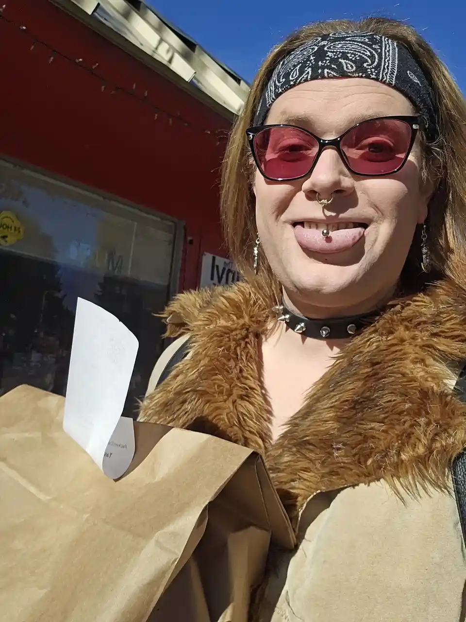 A person with a black bandana, pink sunglasses, a nose ring, and a lip piercing sticks their tongue out while holding a…