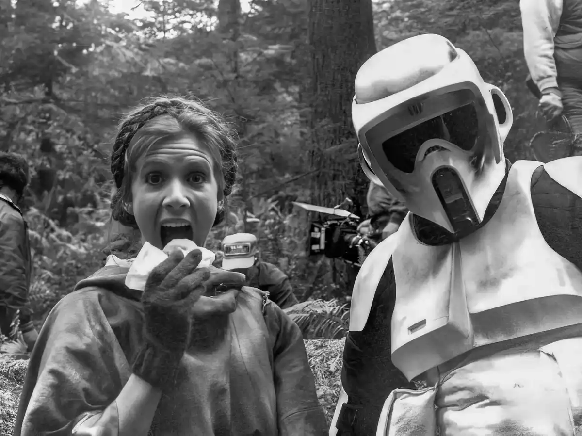 A black-and-white behind-the-scenes photo shows actress Carrie Fisher as Princess Leia, mid-bite with a shocked expression…