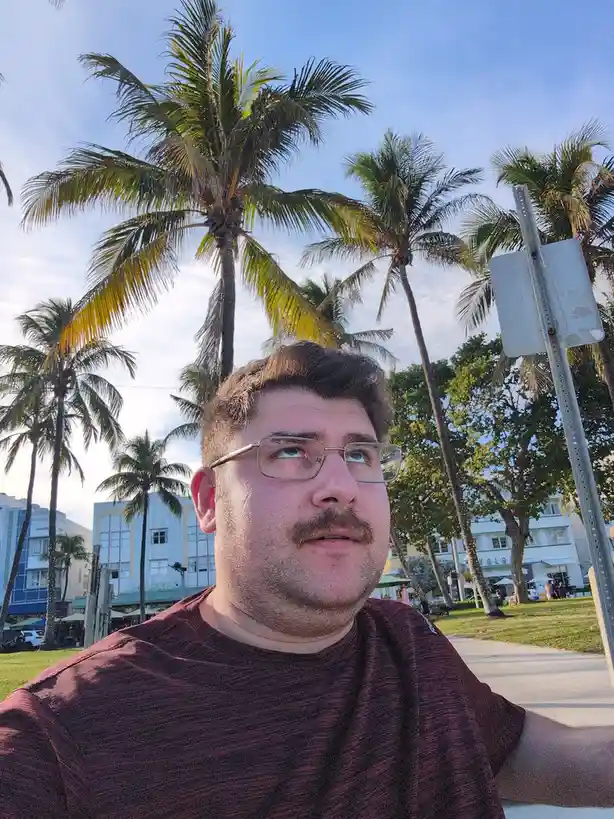 A man with glasses and a mustache looks upward with a slightly confused or distracted expression, standing outdoors in front…