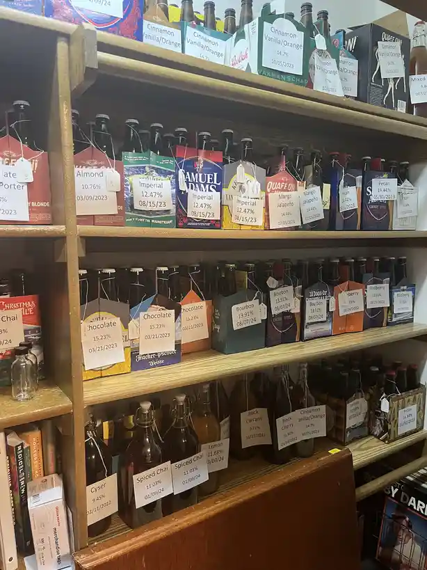 A wooden shelf displays multiple rows of beer bottles and boxes, each tagged with handwritten labels showing flavor names…