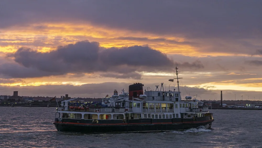 A large, white and black ferry with a red stripe sails on water under a dramatic sunset sky with orange and purple clouds…