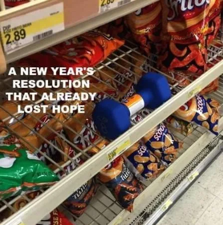 A blue dumbbell sits on a supermarket shelf among bags of chips and snacks, with price tags visible. White text overlaid on…