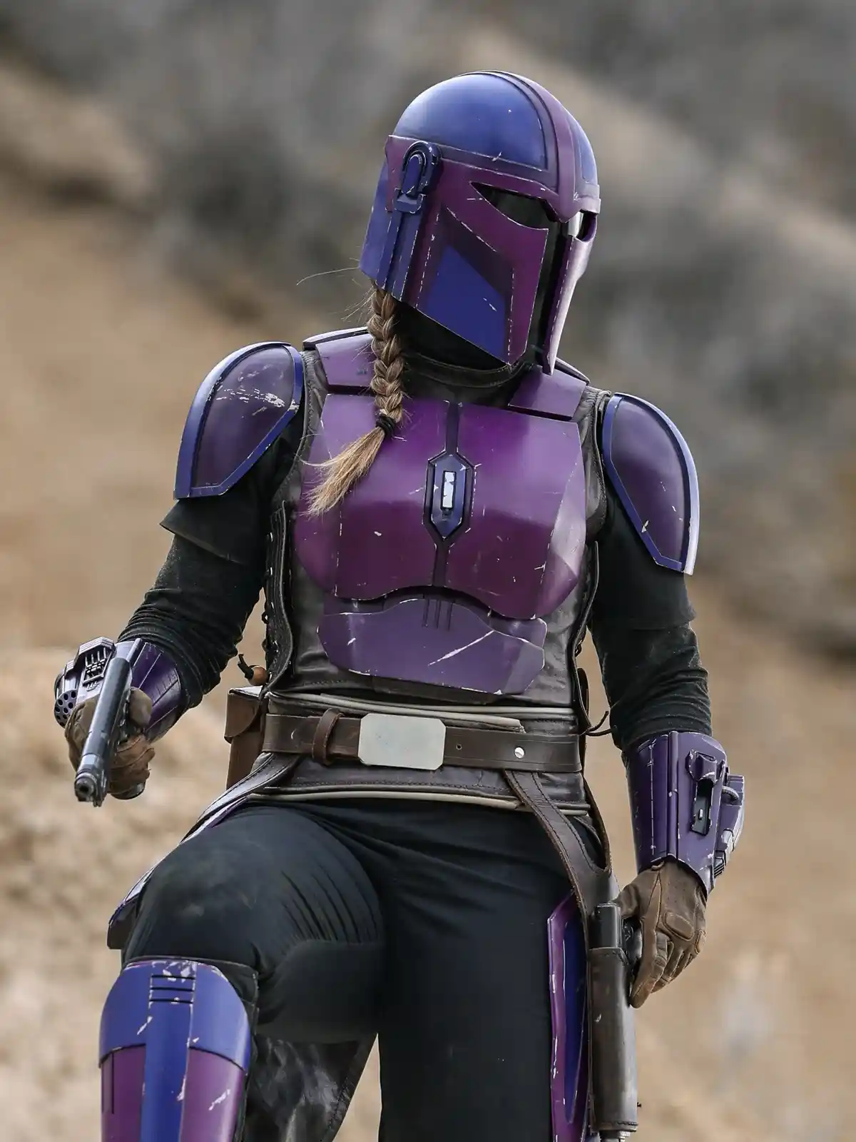 A person in purple and blue Mandalorian armor with a braided ponytail visible under the helmet, holding a blaster pistol in…
