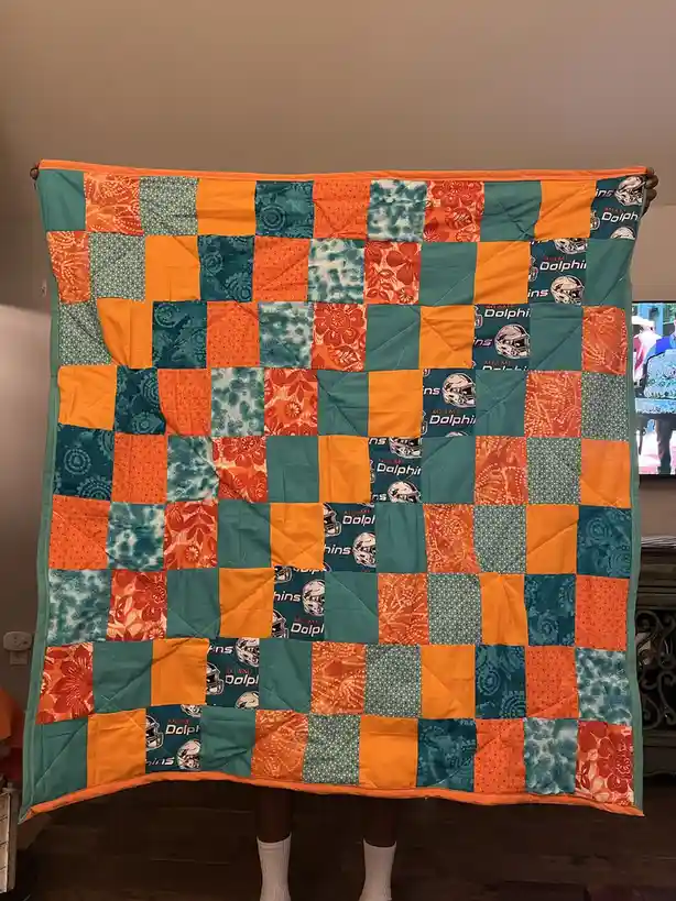 A person holds up a handmade quilt made of orange and teal squares, some featuring Miami Dolphins logos and others with…