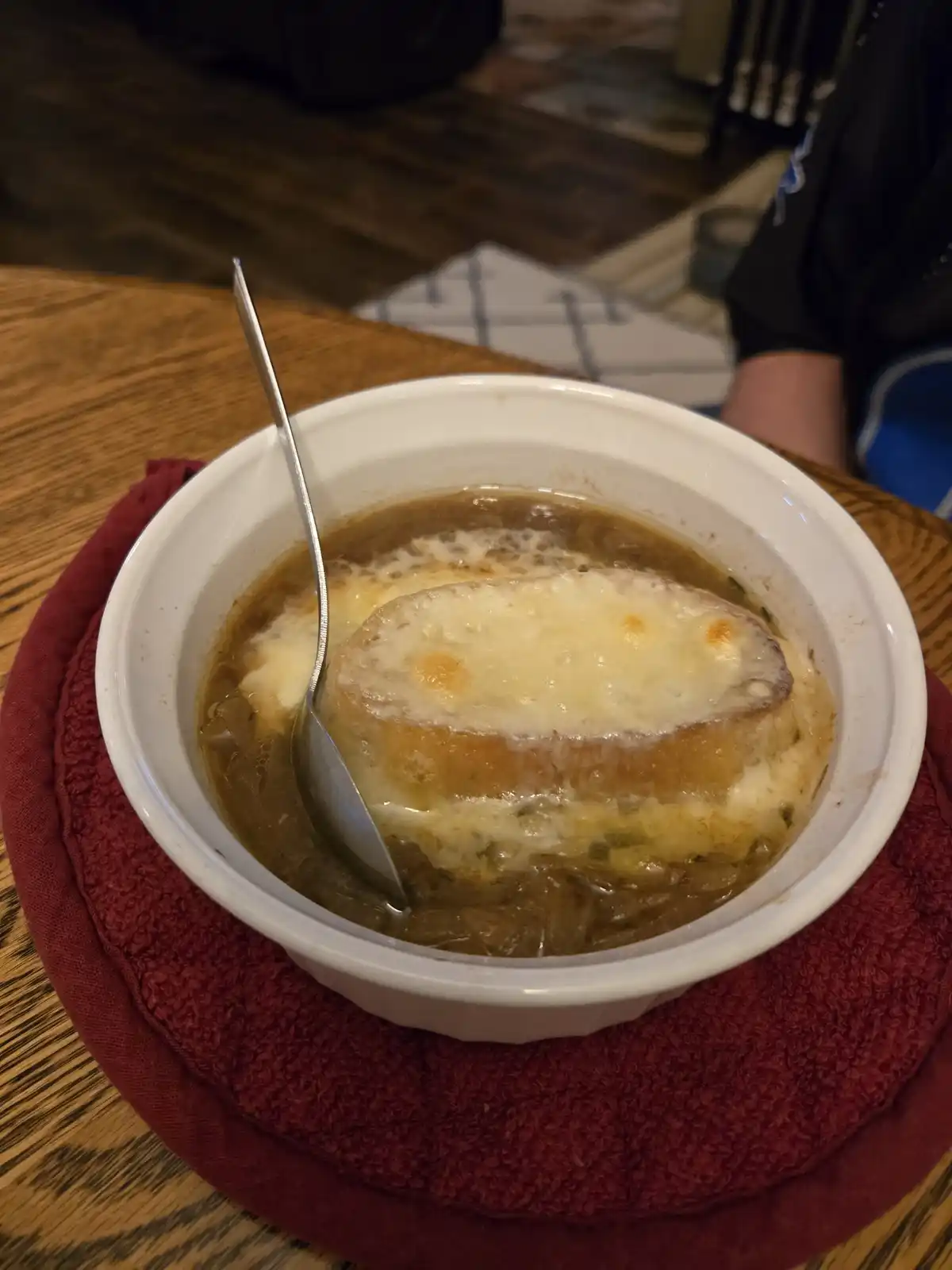 A close-up photo shows a white bowl of French onion soup on a red cloth napkin, resting on a wooden table. The soup has a…
