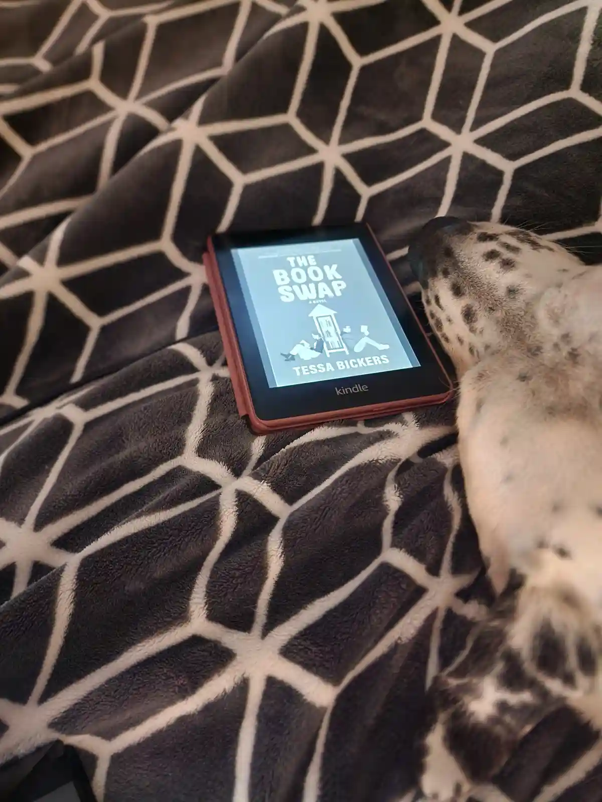 A red Kindle e-reader displaying the cover of 'The Book Swap' by Tessa Bickers rests on a black and white…