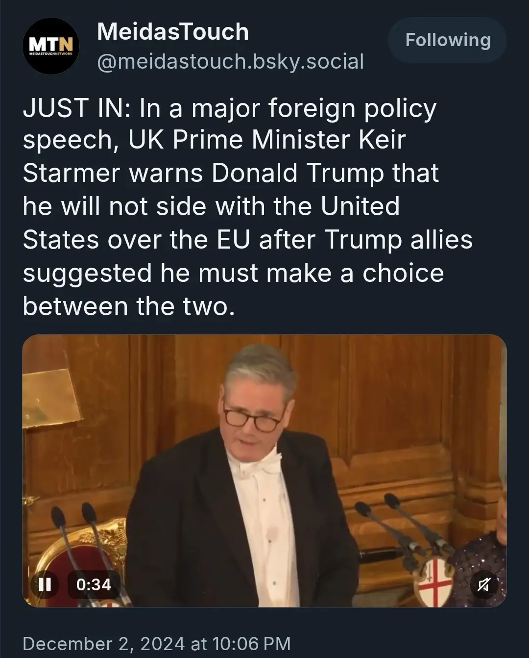 A screenshot of a social media post showing a man in a tuxedo speaking at a podium with microphones, overlaid with text…