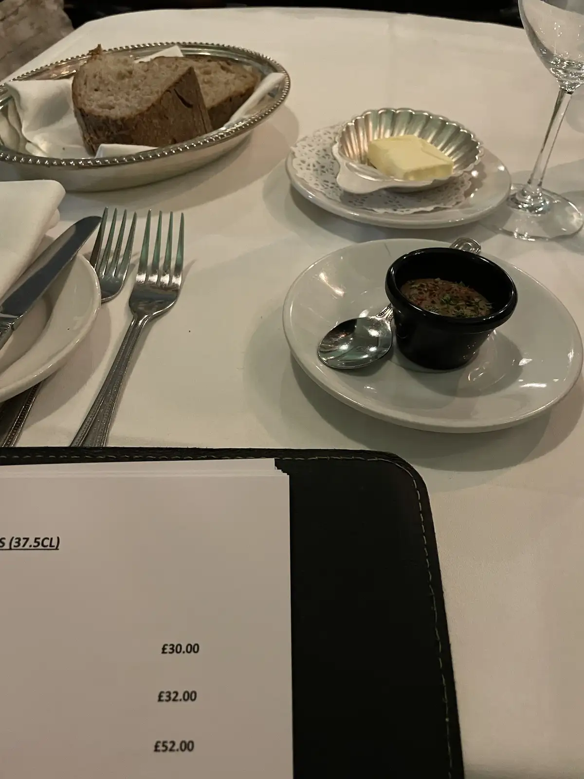 A close-up of a restaurant table setting with bread, butter, and a small black bowl of condiment, with a menu in the…