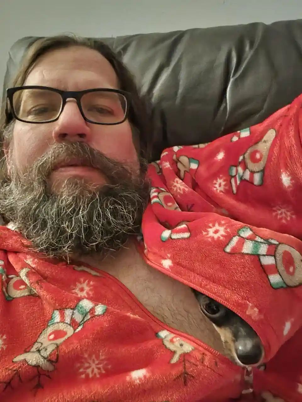 A man with a full beard and glasses lies on a dark couch, wrapped in a red Christmas-themed blanket featuring reindeer and…