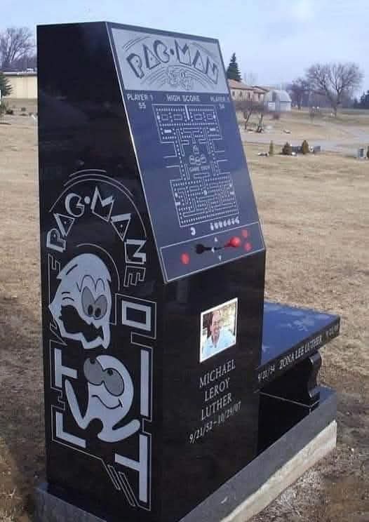 A black tombstone shaped like a Pac-Man arcade machine, featuring the game's logo, maze, and character. It includes a small…