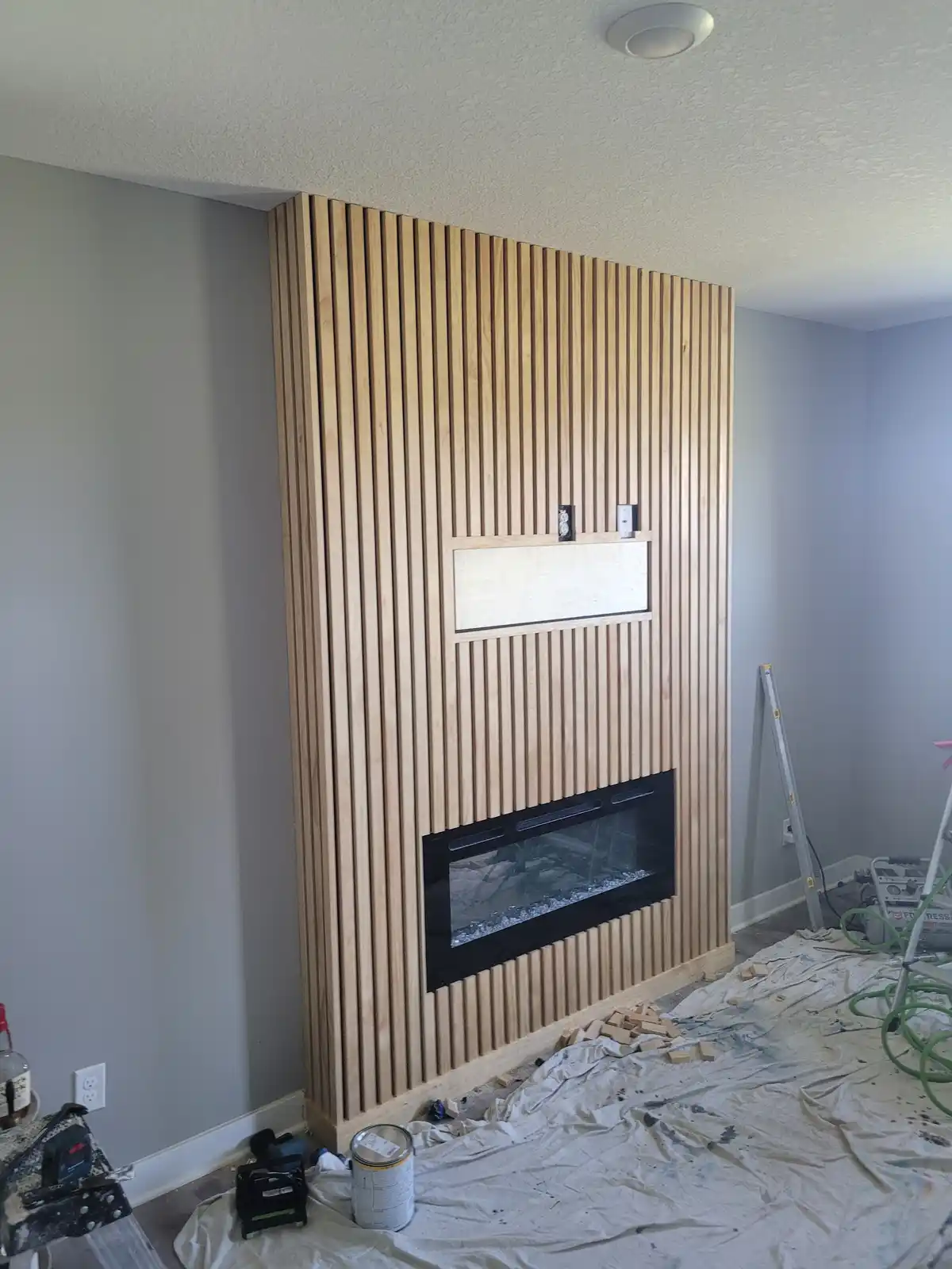 A room under renovation shows a newly installed wooden slat fireplace wall with a black electric fireplace insert and a…