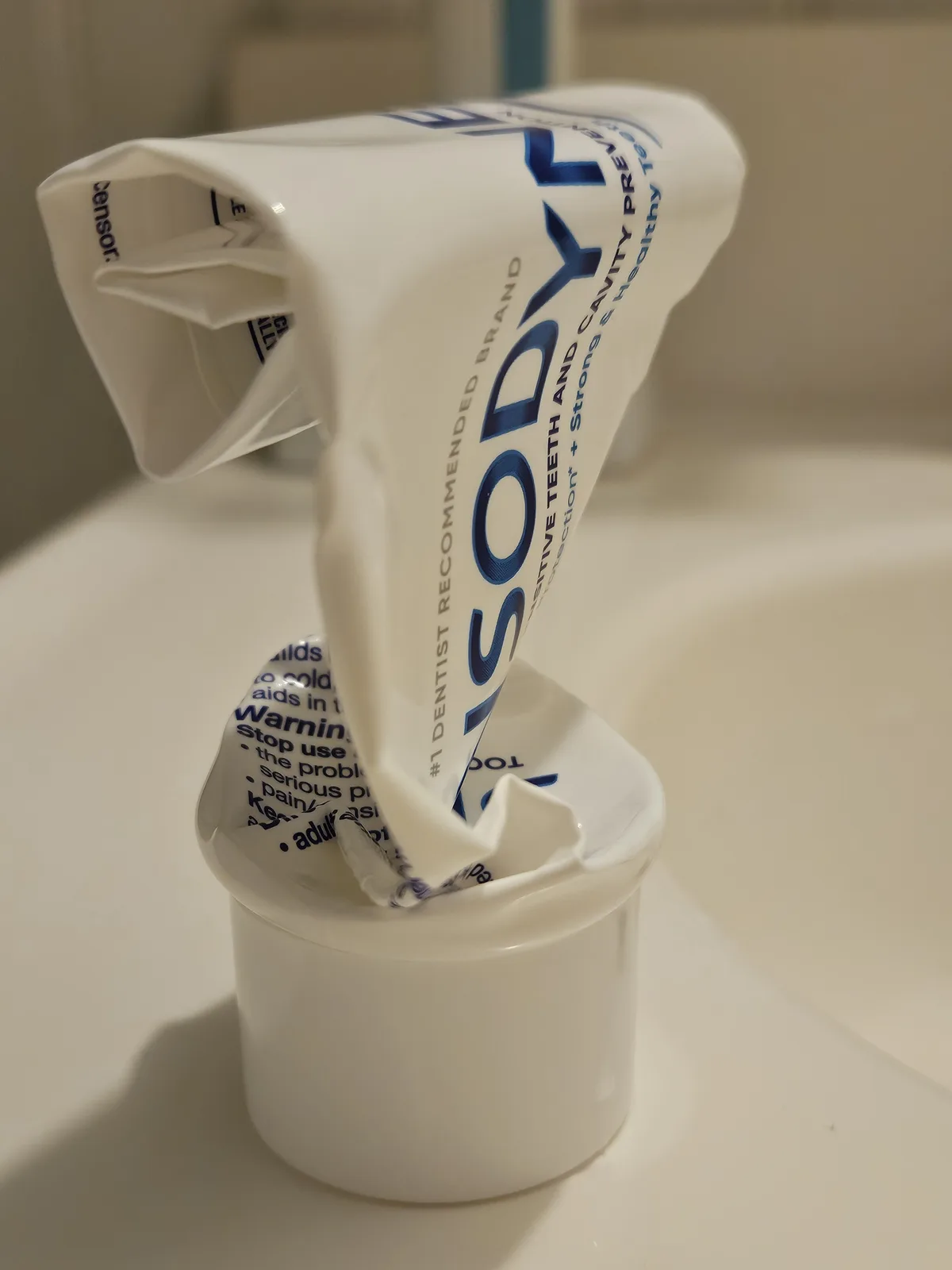 A white tube of Sensodyne toothpaste is visibly squeezed and inverted, with its contents spilling out into a small white…