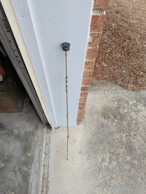 A rusty bolt protrudes vertically from a white-painted wooden door frame, extending down to the concrete ground. The bolt is…