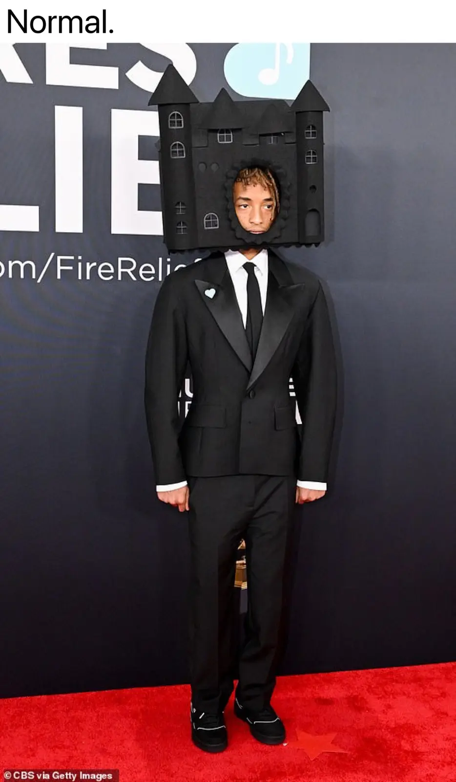 A person in a black tuxedo stands on a red carpet with a large black castle structure covering their head, with the word…