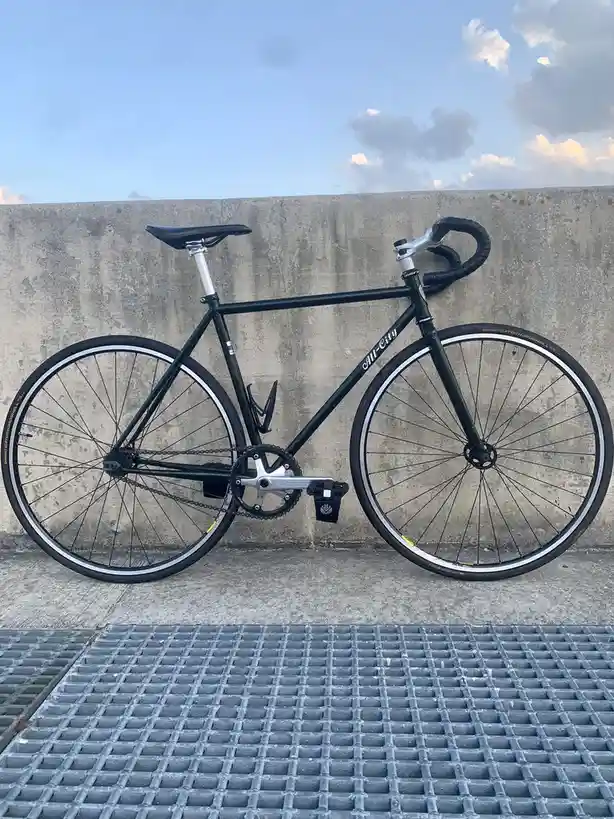 A black fixed-gear bicycle with 'All-City' branding on the frame leans against a weathered concrete wall under a partly…