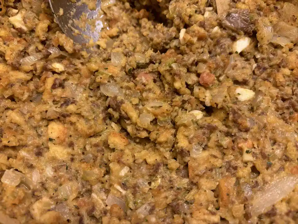 A close-up photo of golden-brown, textured stuffing with visible pieces of bread, onions, herbs, and meat, being stirred…