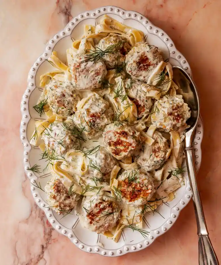 An overhead shot of a white, scalloped-edge platter filled with creamy pasta topped with numerous meatballs and garnished…