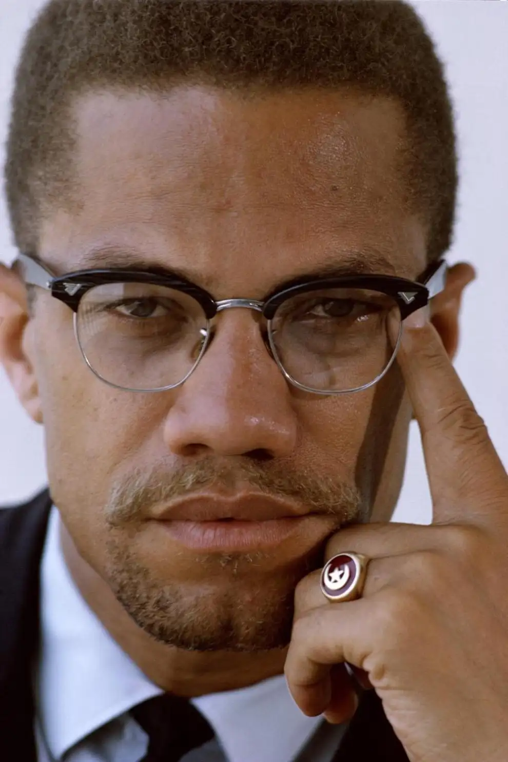 A close-up portrait of Malcolm X wearing glasses and a suit, with his hand resting near his face and a distinctive ring on…