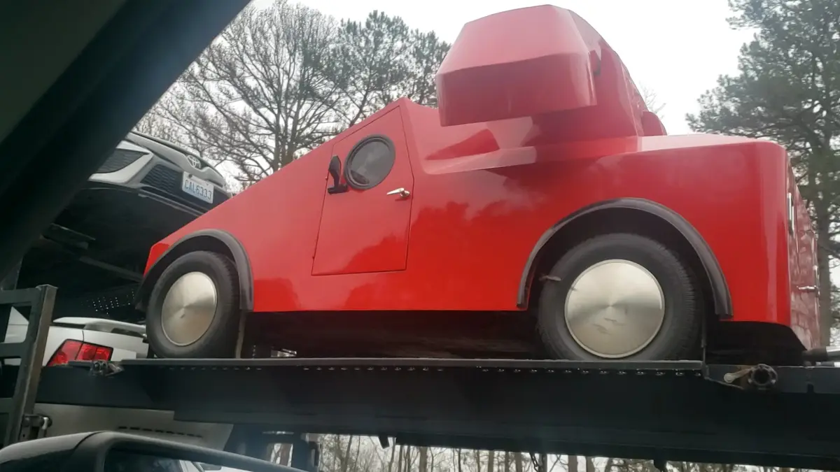 A bright red, angular, custom-built vehicle with a boxy shape, round window, and silver hubcaps is loaded onto a black car…