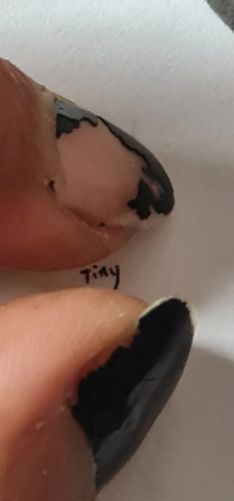 A close-up photo shows two fingers with chipped black nail polish against a white background. The word 'Tiny' is handwritten…