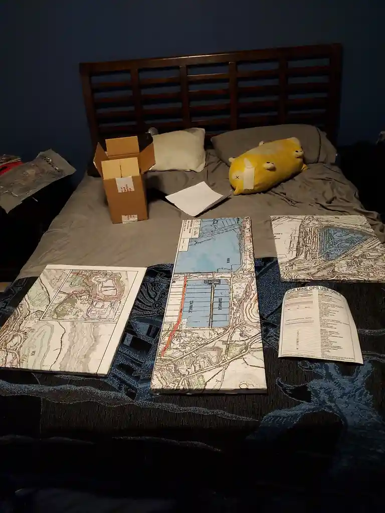 A bed is covered with topographic maps, a cardboard box, and a yellow plushie, with text overlaid that reads 'FOR MINE OPS…
