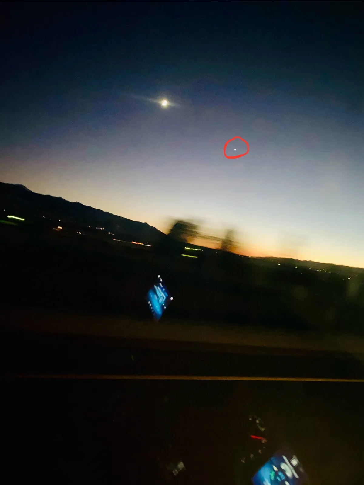 A blurry nighttime photo taken from inside a moving car shows a dark sky with a bright moon and a small, faint light circled…