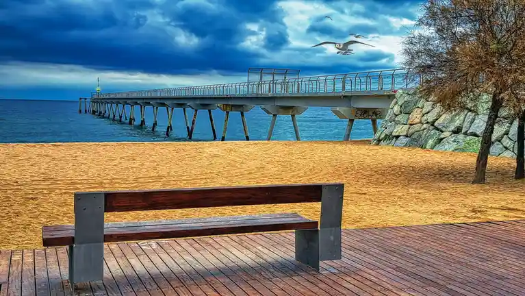 A wooden bench sits on a wooden boardwalk overlooking a sandy beach and a long pier extending into the ocean under a…