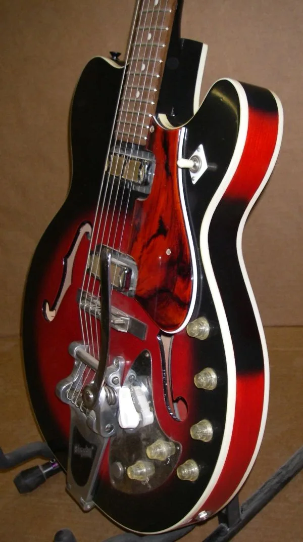 A close-up photo of a red and black semi-hollow body electric guitar with a white binding, showing a visibly broken bridge…