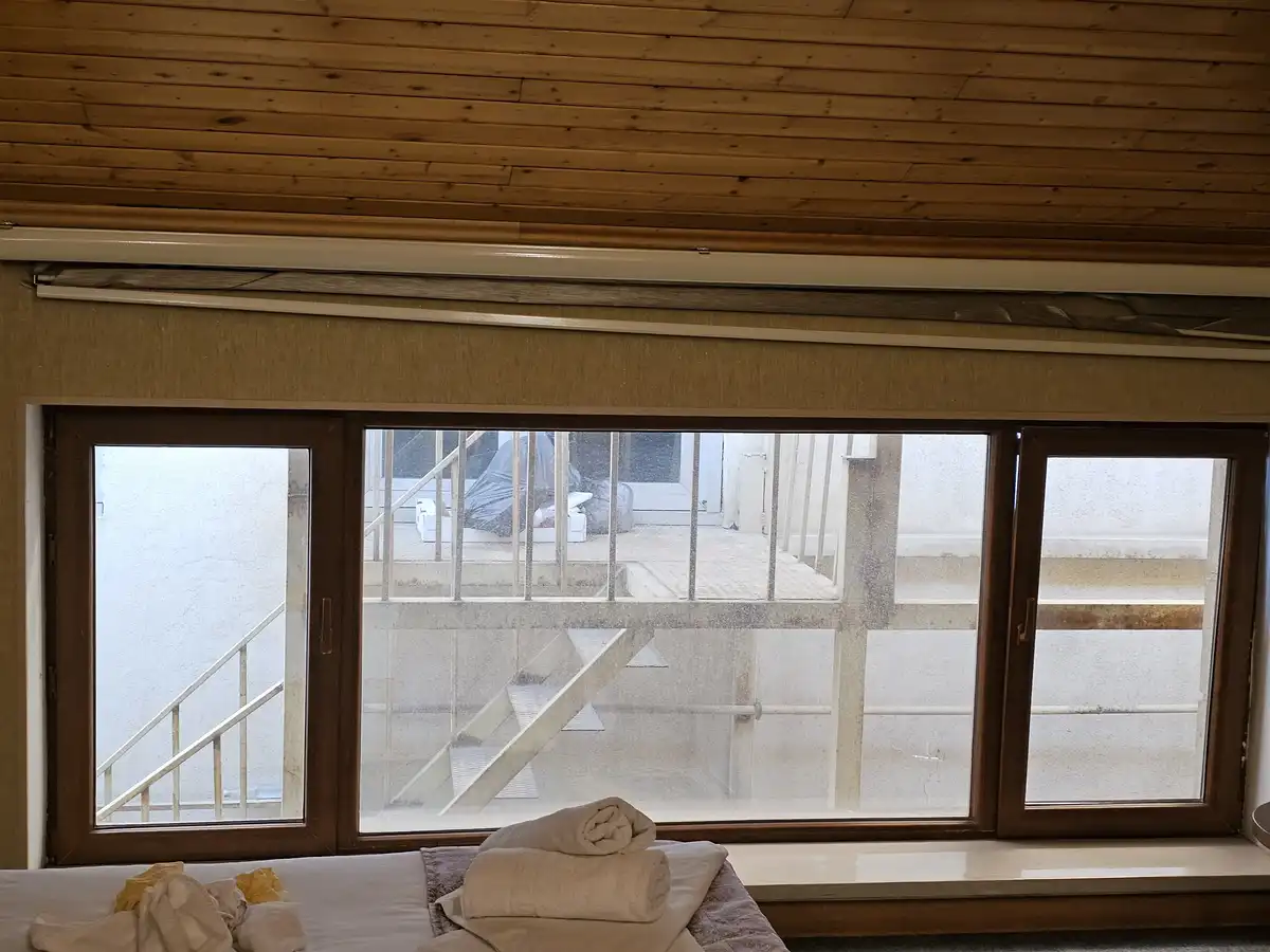 A photo shows a bedroom with a wooden ceiling and a large window looking out onto a concrete balcony with stairs. Rolled…